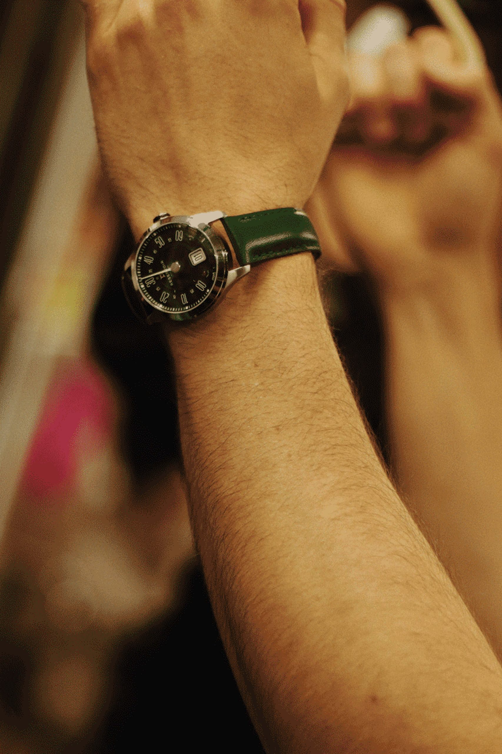 Bras d'une personne portant une montre-bracelet avec cadran noir et cuir vert bracelet, fond flou.