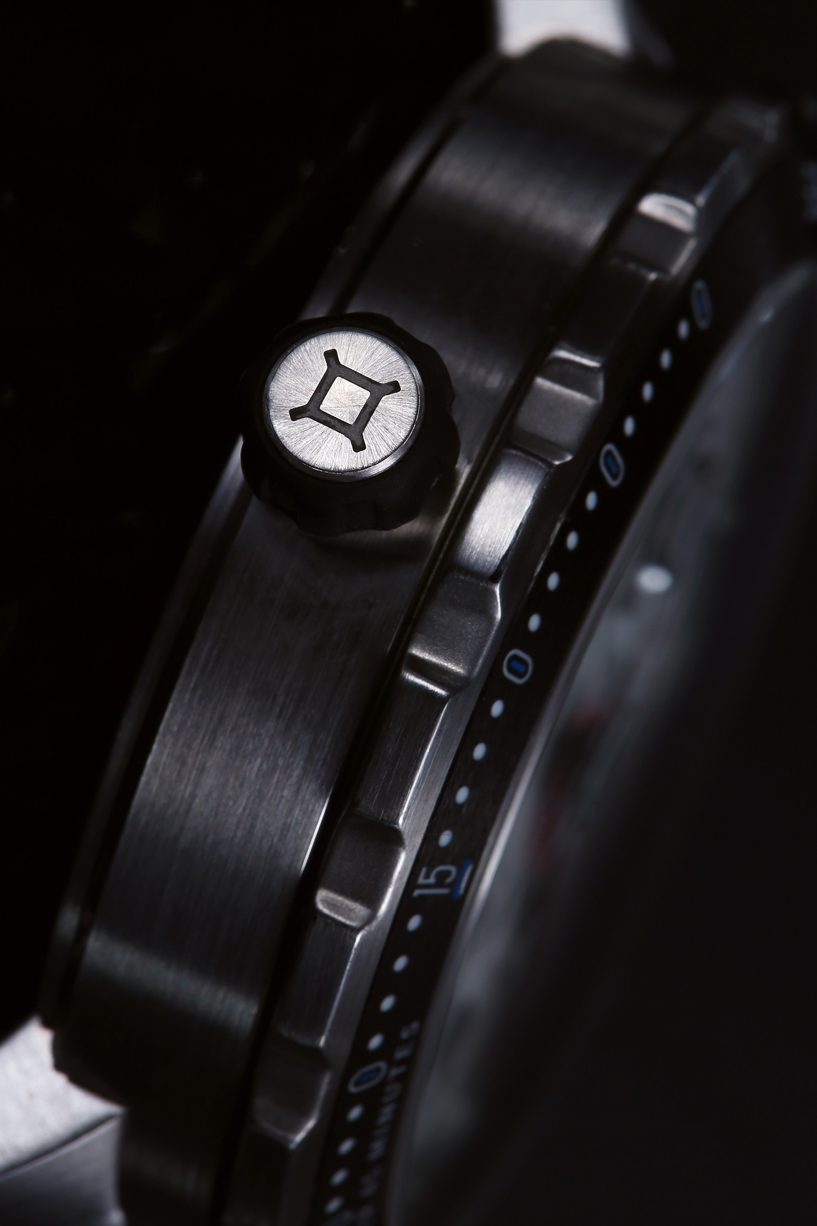 Close-up of a wristwatch side showing a metallic finish, crown with geometric design, and minute markings.