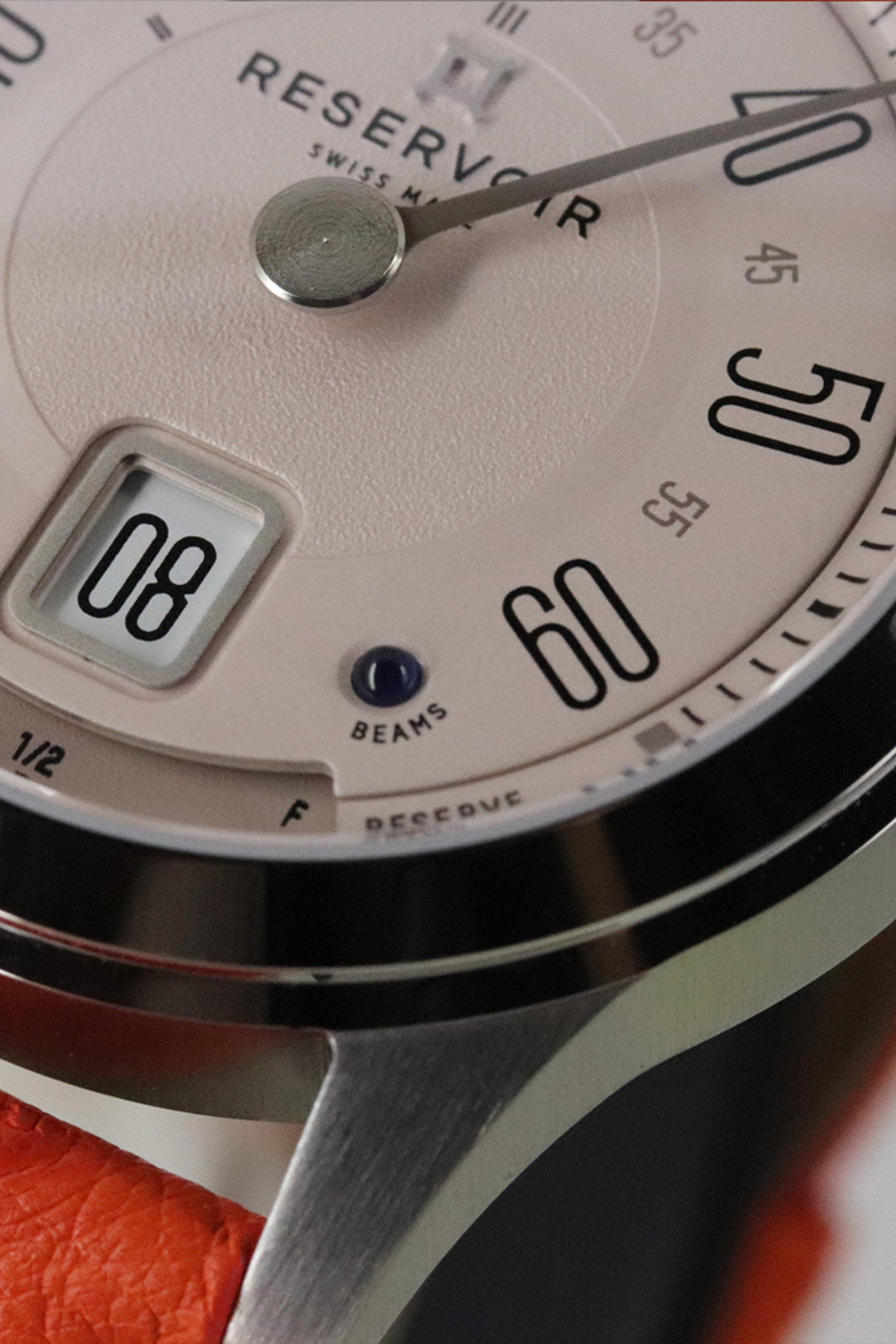 Gros plan sur une montre-bracelet avec cadran beige, chiffres noirs, date "08", bracelet rouge-orange, marque "RESERVOIR".