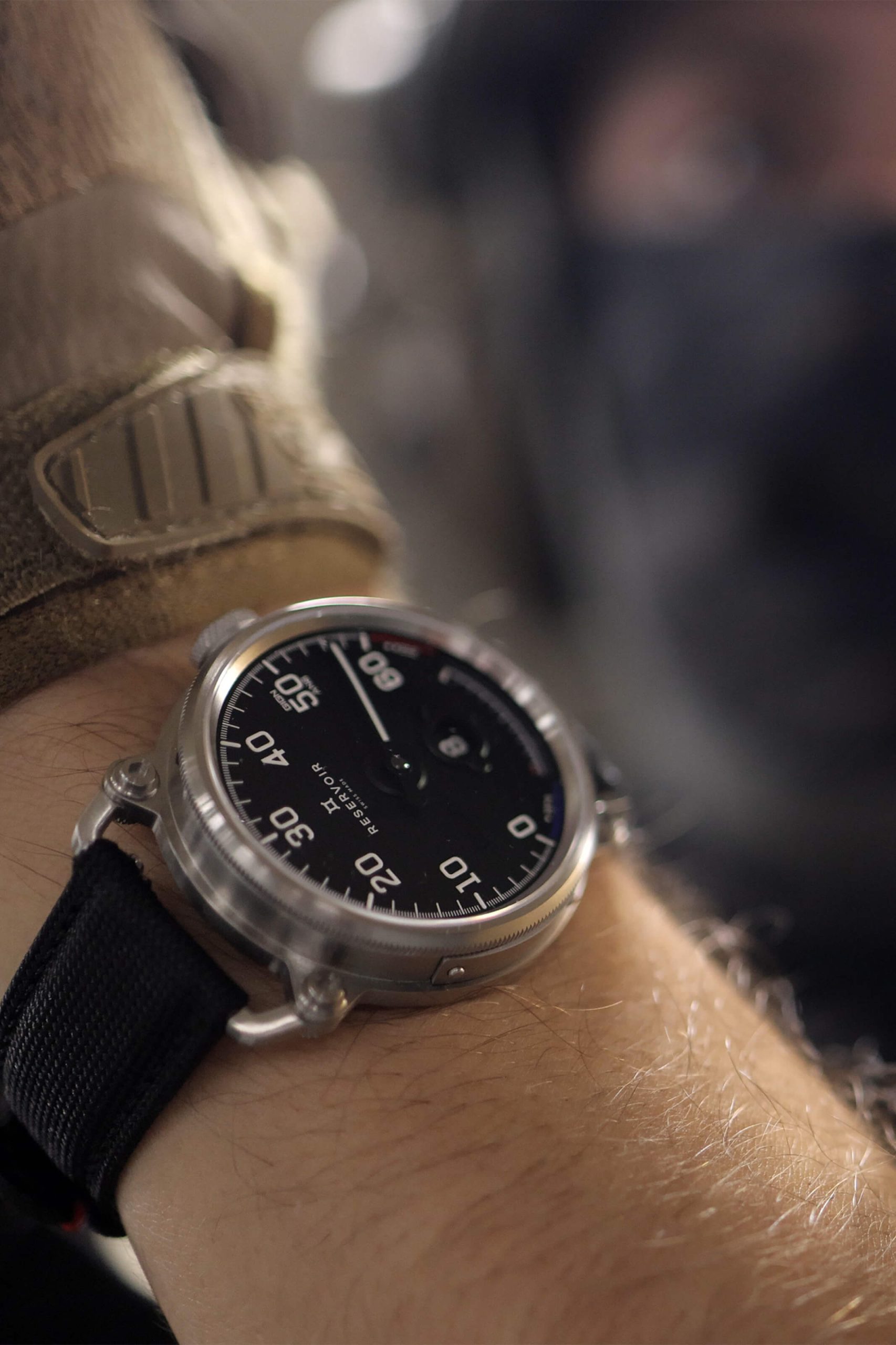 Un poignet portant une montre ronde à bracelet noir avec des chiffres en gras ; silhouette floue en tenue de camouflage.