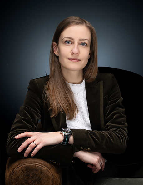 Femme aux longs cheveux bruns, vêtue d'un blazer foncé et d'un haut en dentelle blanche, assise, les bras croisés.