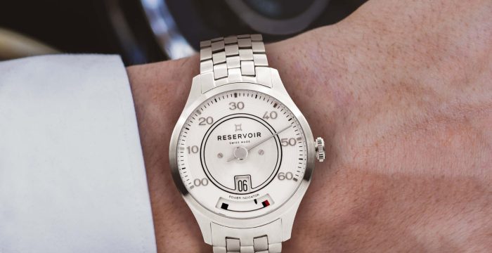 Close-up of a Reservoir watch on a wrist, white dial, metal band, in front of a wooden steering wheel.