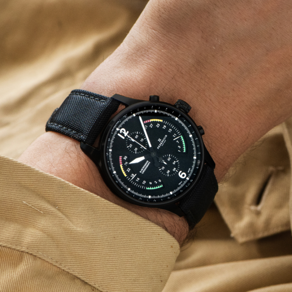 Close-up of a wrist wearing a black watch with subdials, black fabric strap, person in light brown garment.