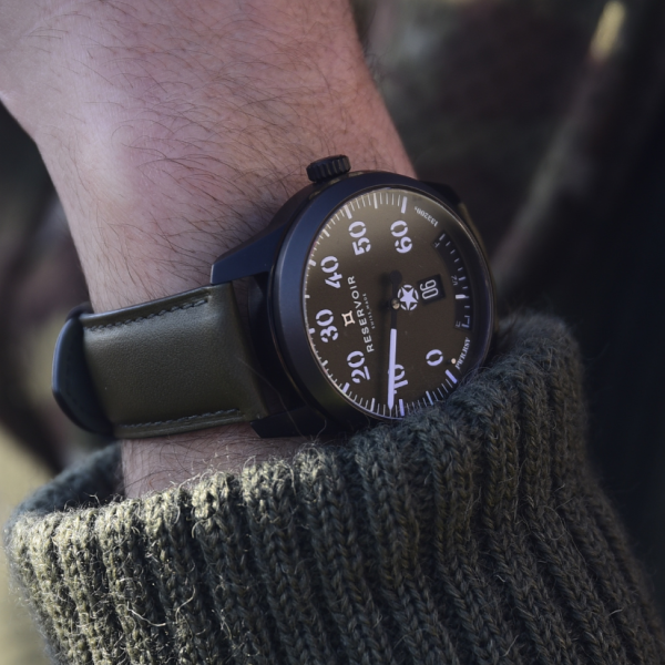 A wristwatch with a dark dial, white numerals, date display, black case, and olive green leather strap.
