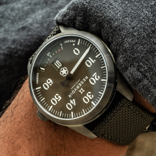 Close-up of wristwatch with black dial, bold white numerals, canvas strap, worn on grey fabric sleeve.