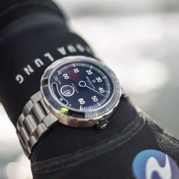 Wristwatch with metallic bracelet and gauge-like dark face on black sleeve with white and blue logos.