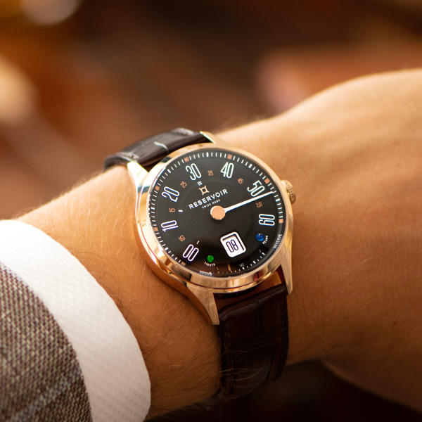 Wristwatch with black dial, gold casing, brown leather strap, worn with white shirt and jacket.