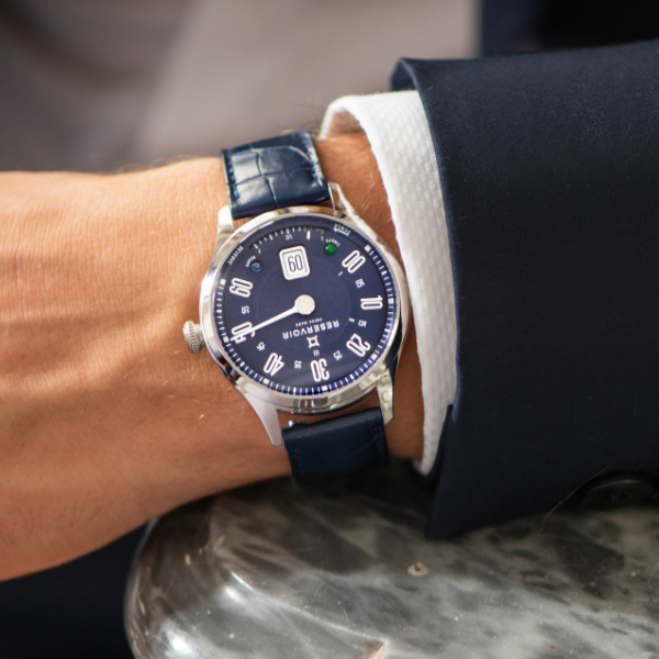 Person wearing a blue-dial wristwatch with white numerals, blue strap, and dark suit, marble background.