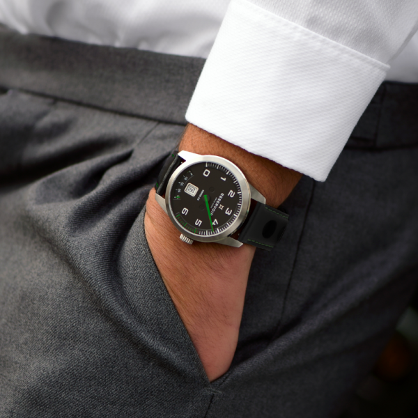 Person in a white shirt, grey trousers, wearing a black wristwatch with green accents.