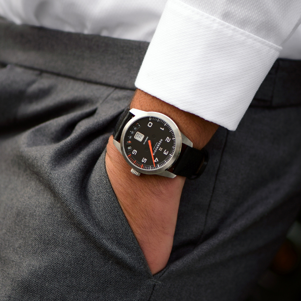 Hand in pocket wearing grey trousers, white shirt, and black wristwatch with Arabic numerals.