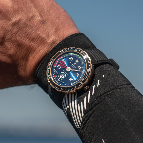 Wrist wearing a black strap wristwatch with a multi-colored dial, against a clear blue sky background.