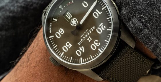 Wrist wearing a black analog watch with large white numerals and a black textured strap.