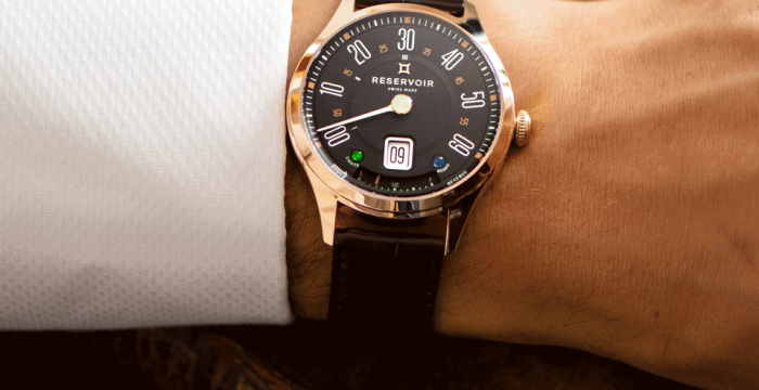 Wrist wearing a RESERVOIR watch with black dial, white numerals, leather strap, near old book.