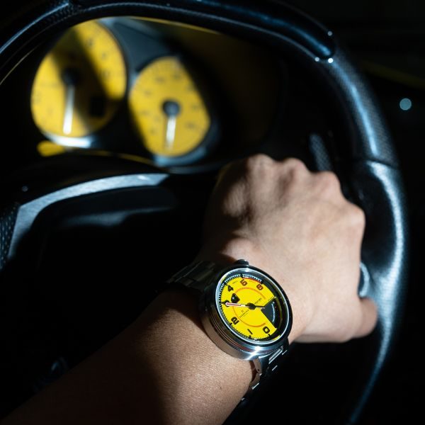 Hand with yellow-faced wristwatch gripping steering wheel; yellow gauges in a dimly lit car dashboard.