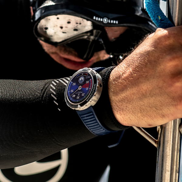 Person in diving suit and goggles, holding metal railing, smartwatch with blue strap showing dive interface.