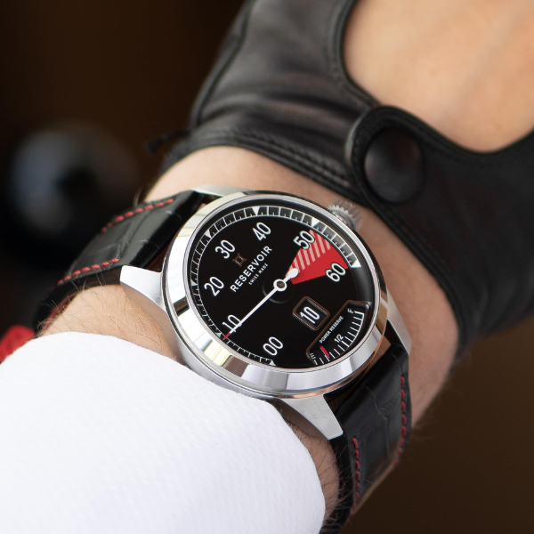 Wristwatch with black dial, white numerals, red minute indicator, black strap, silver casing, worn with glove.