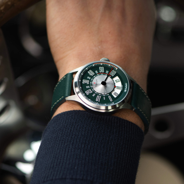 Wristwatch with green dial, white markers, red accents, green strap, worn with dark blue sleeve.