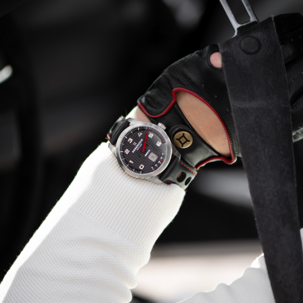 Person gripping a strap, wearing a black leather glove and wristwatch with a black dial and red details.