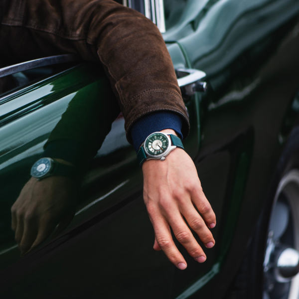 Close-up of an arm resting on green car door, wearing brown jacket, blue sleeve, and green wristwatch.