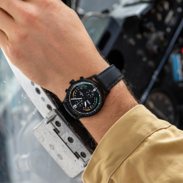 Wrist with black analog watch, multiple dials, resting on metal surface in aircraft cockpit. Beige shirt.
