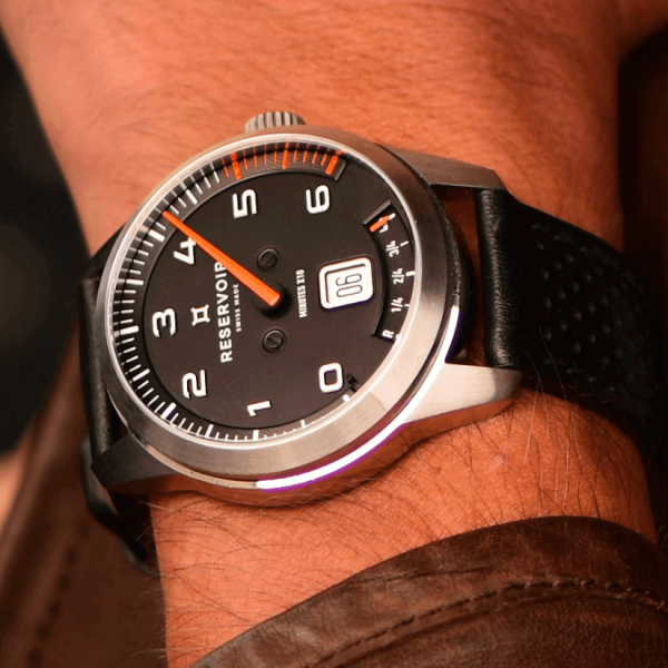 Wristwatch with black face, white numerals, orange minute hand, date at 3, black textured strap.