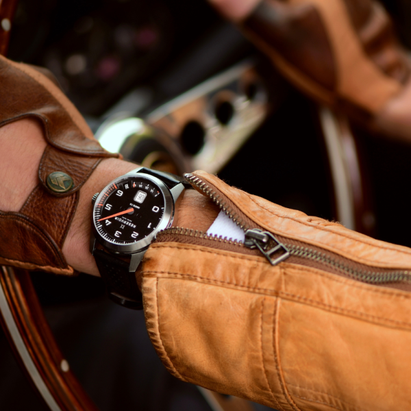 Individual wearing a black analog watch, brown leather gloves, and a tan jacket with a visible zipper.