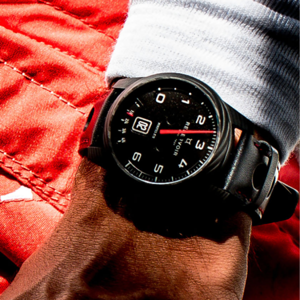 Black wristwatch with white numerals and red second hand on wrist, red fabric background.