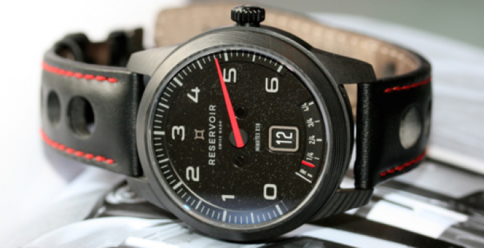 Black wristwatch with white numerals, red-stitched strap, and "RESERVOIR" brand on an open book.