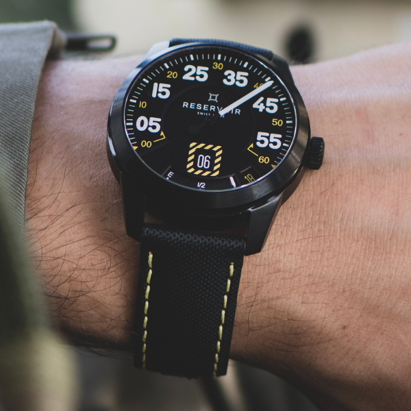 Black wristwatch with white numerals, date display, branded "Reservoir," black strap with yellow stitching.