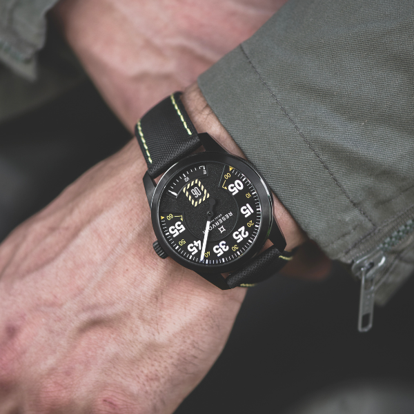 Black wristwatch with yellow accents, black strap with green stitching, worn with gray-green jacket.