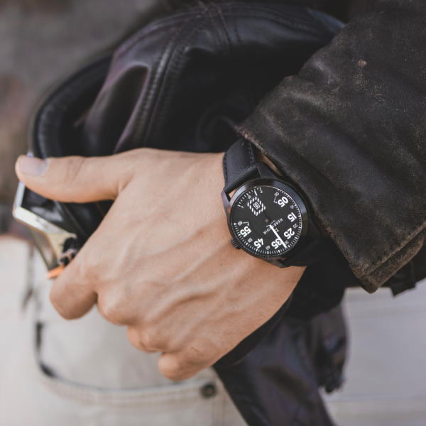 Person wearing a black watch with white numbers, holding a black leather jacket, dressed casually.