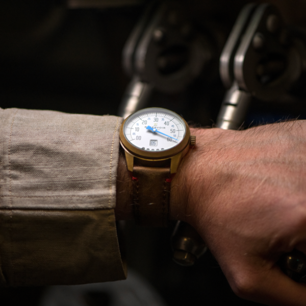 Close-up of a wrist wearing a gold-case watch with white dial, blue hands, date, and brown leather strap.