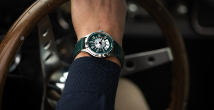 Hand wearing green wristwatch gripping wooden steering wheel, dark sleeve, blurred vehicle interior.