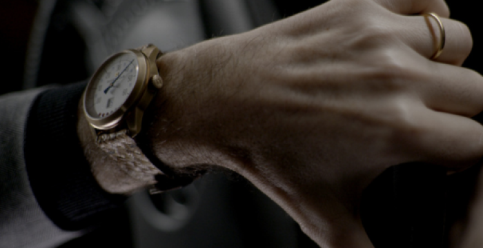 Hand wearing a gold ring and wristwatch with a brown strap, gripping a steering wheel.