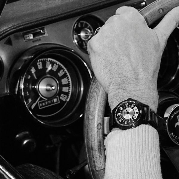 Hand gripping vintage car steering wheel, wristwatch visible, analog dashboard gauges in background