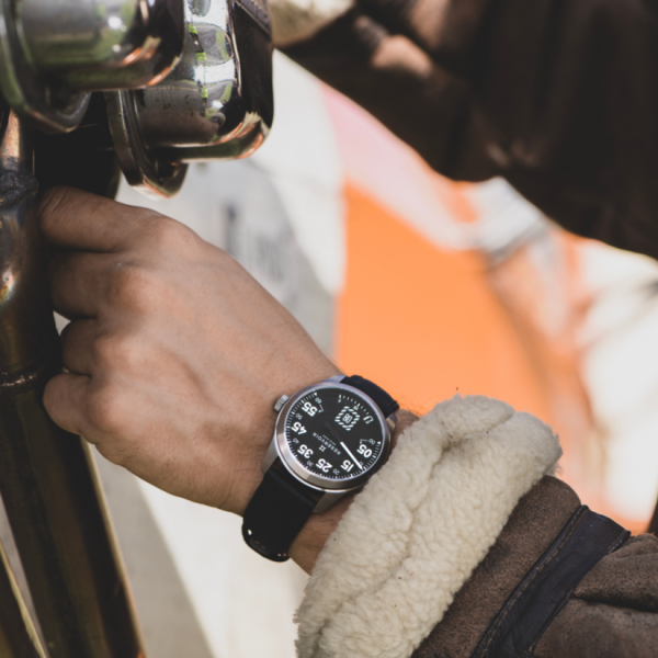 Hand wearing a wristwatch with black strap, holding a metallic item; brown jacket with shearling.