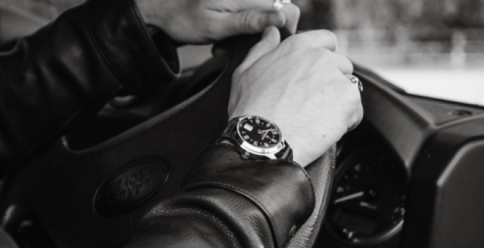 Person wearing a leather jacket and wristwatch gripping a steering wheel with blurred background.