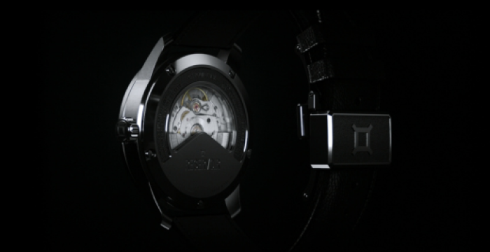 Luxury mechanical wristwatch with visible gears through transparent back; black strap and distinctive clasp.