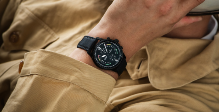 Person wearing beige jacket showcasing a black chronograph wristwatch with detailed dial and sub-dials.