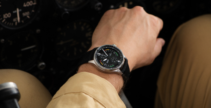 Person in cockpit wearing a black strap wristwatch with a round, black face displaying various dials.