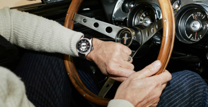 Person driving a vehicle with a wooden steering wheel, wearing a watch and light-colored sweater.