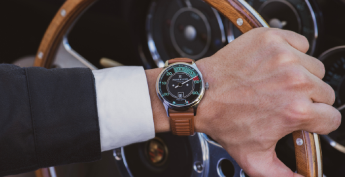 A person in a dark suit holds a wooden steering wheel, showing a brown leather-strap wristwatch.
