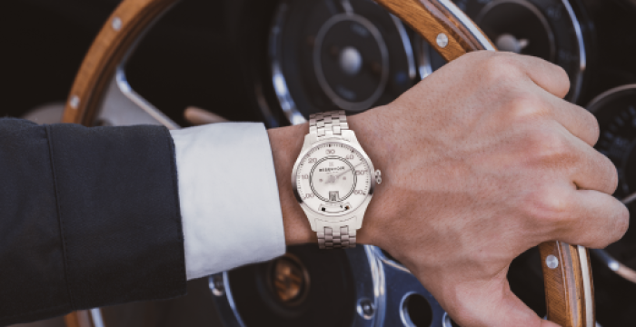 Person wearing silver wristwatch holds wooden-stripped steering wheel in car with visible dashboard.