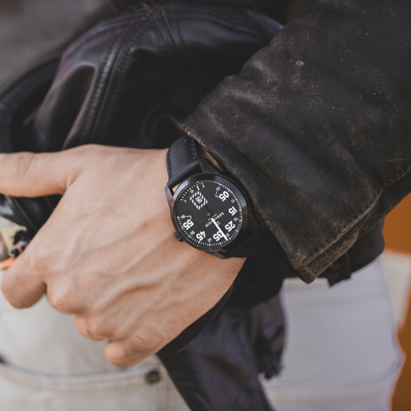 Person's wrist with black watch, white numerals, holding black leather jacket, wearing light pants.