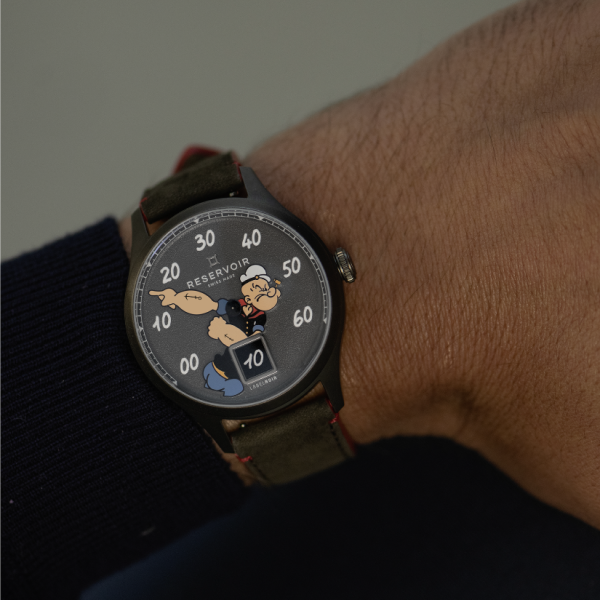 Close-up of a wristwatch with Popeye on the dial, dark case, leather strap, and RESERVOIR branding.