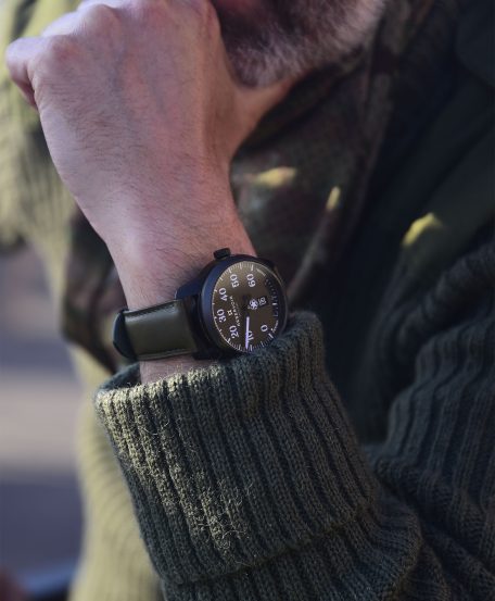 Person in green sweater, camouflage scarf, resting chin on hand, wearing black watch with green band.