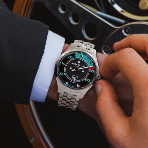 Person adjusting a Reservoir wristwatch with black dial, on wrist with dark suit in metallic background.