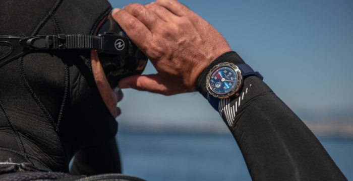 Person in a black wetsuit adjusting scuba mask, with a blue watch, near the water and sunny sky.