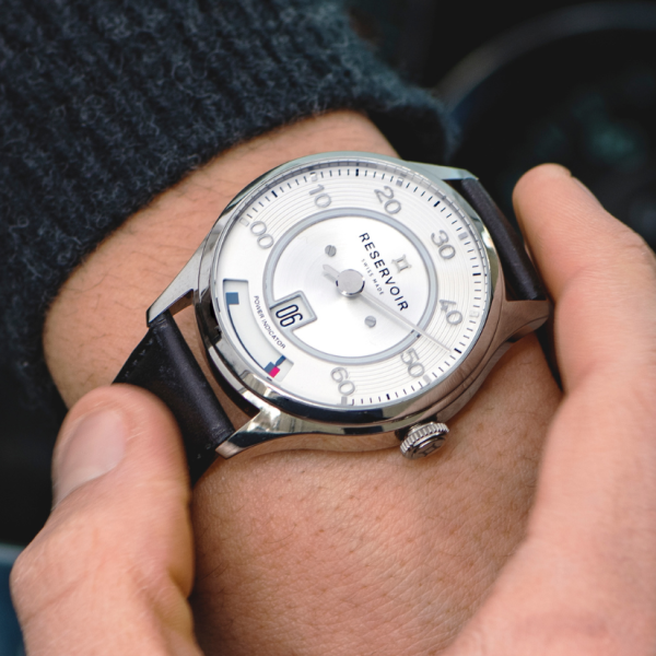 Wrist wearing a silver analog watch "Reservoir" with a white dial, black leather strap, date "06".
