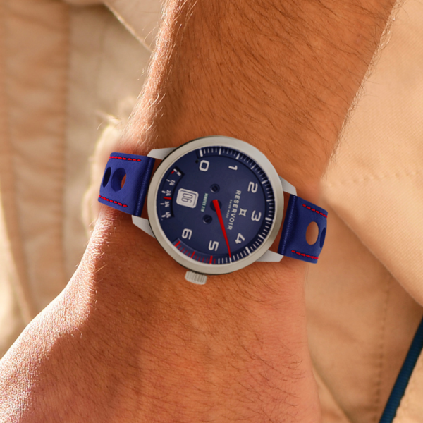 Wrist wearing a stainless steel watch with blue dial, blue strap, white numerals, and date window.